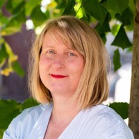 Portrait of a smiling blonde woman wearing a light blue blouse, outdoors among green leaves.