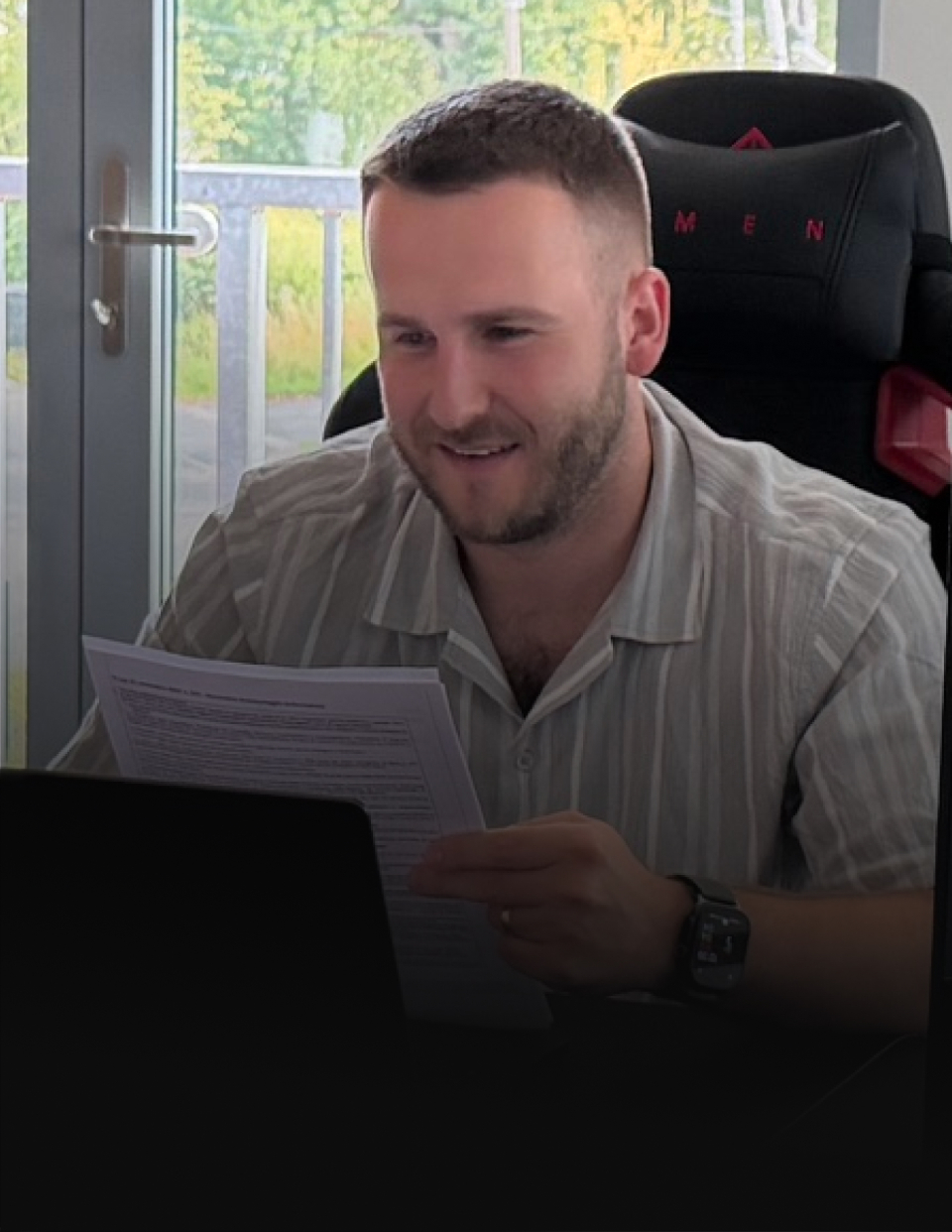 Man with short hair and beard sits at a desk, reading a document in front of a laptop near a glass door window.