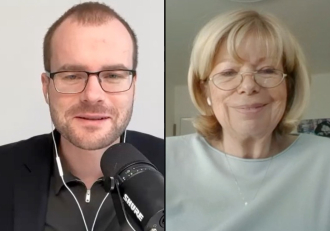 Split-screen video call: man on the left with glasses speaking into a microphone, wearing a black blazer; woman on the right smiles, wearing glasses.