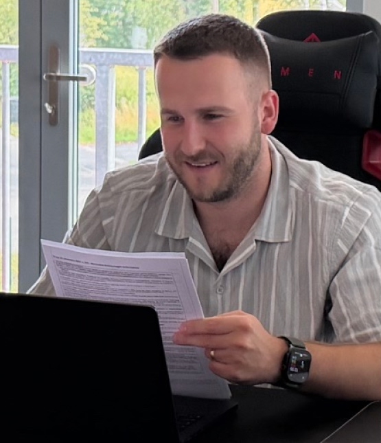 Man with short hair and beard in a striped shirt sits at a desk, reading printed documents beside a laptop.