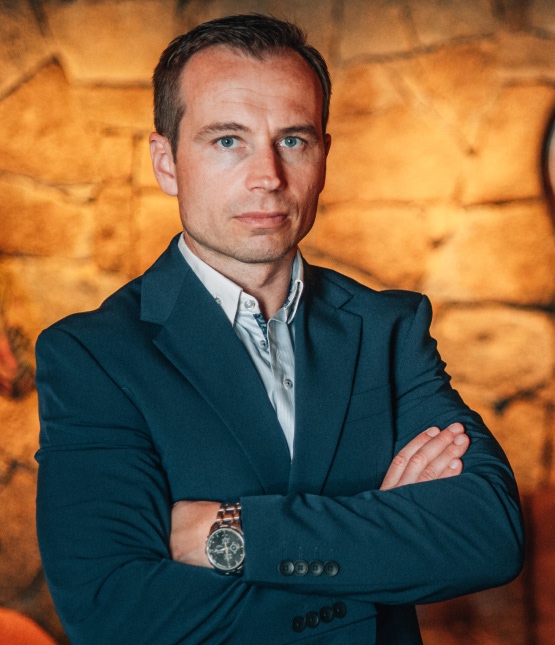 Man in a blue blazer with arms crossed, standing against a warm brick wall.