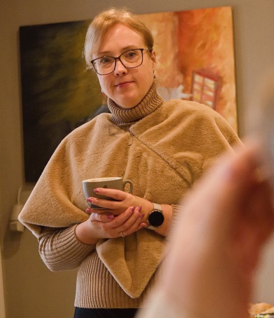 Woman with glasses holding a mug indoors, wearing a beige poncho and turtleneck sweater.