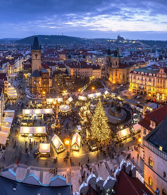 Evening view of Prague's Old Town Square filled with Christmas market stalls, a large decorated tree, and historic buildings nearby.