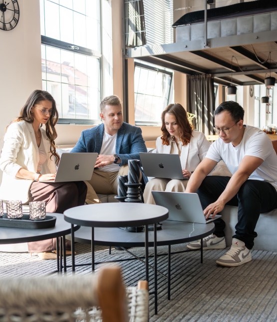 Group of four coworkers seated on a sofa using laptops for a collaborative meeting in a bright loft-style office.