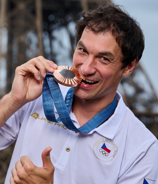 Man in a white polo shirt with a Czech Olympic logo bites a bronze medal and gives a thumbs-up.
