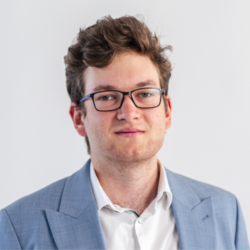 Portrait of a young man wearing black-rimmed glasses and a light blue blazer, looking at the camera.