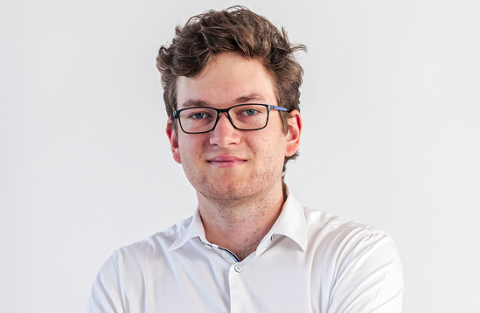 Portrait of a young man with short brown hair and rectangular glasses, wearing a white shirt, looking at the camera.