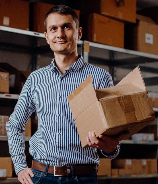 Man wearing a blue striped shirt holds a cardboard box in a warehouse aisle with storage shelves behind him.