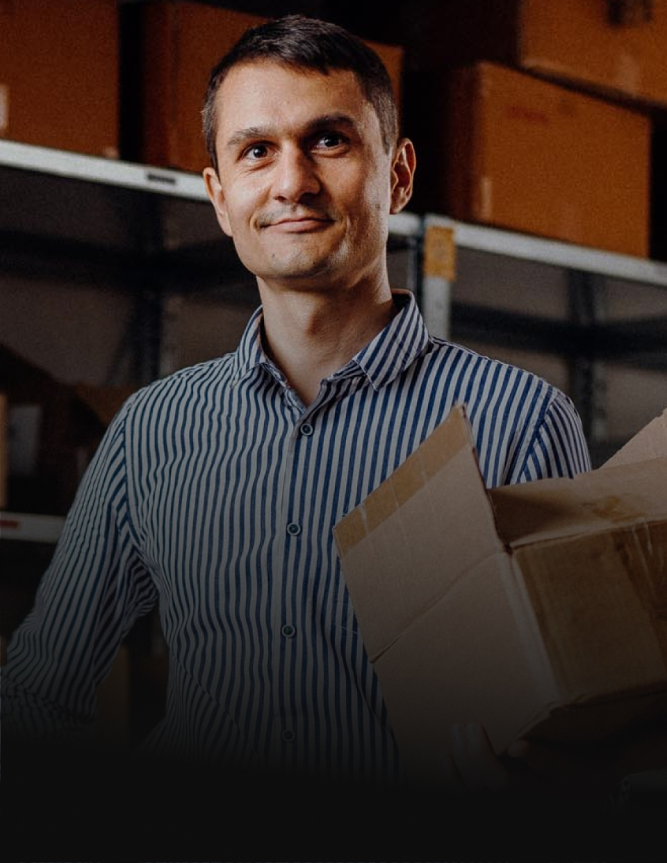 Man in a striped shirt holding a cardboard box in a dim warehouse.