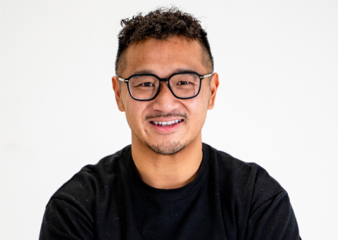 Smiling man with black framed glasses wearing a black shirt against a light background.