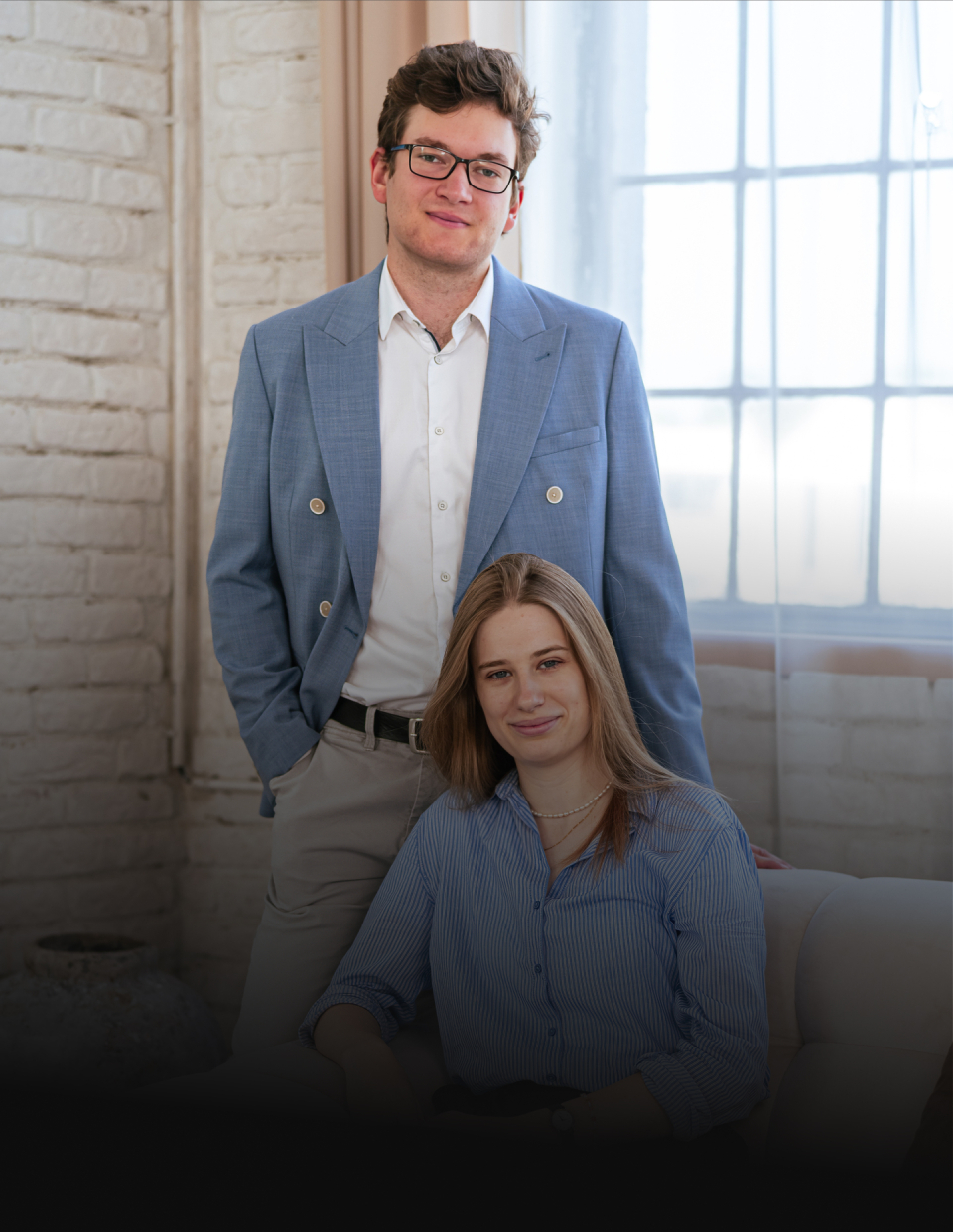 Man in a light-blue blazer stands behind a seated woman in a striped blue shirt, in a bright room with a brick wall and large window.