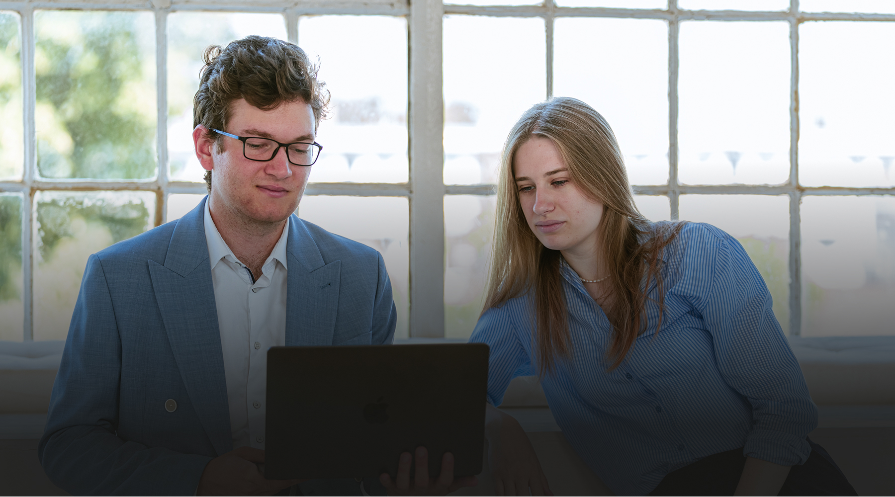 Two young professionals review content on a tablet in a sunlit room by a large grid-window.