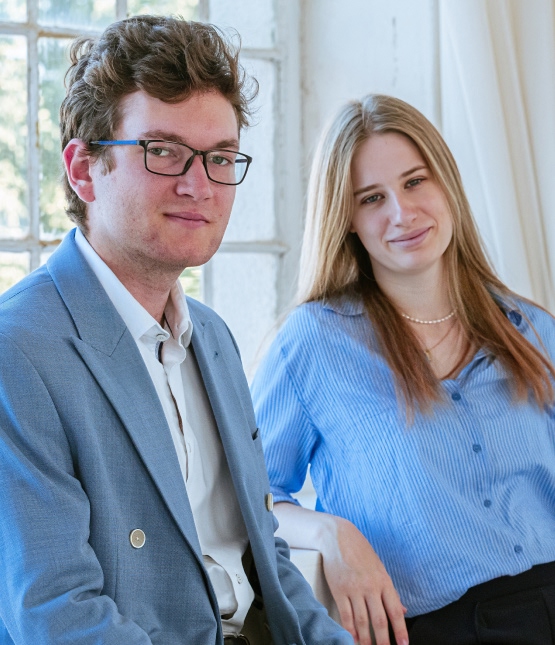 Two young professionals posing for a bright, casual business photo in an office setting, smiling at the camera.