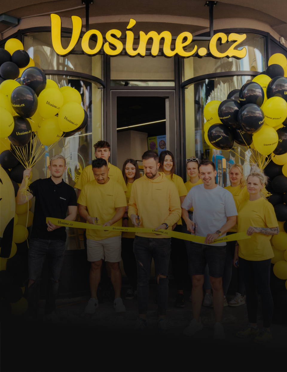 Group of staff in yellow shirts at a store opening, cutting a yellow ribbon beneath a yellow-and-black balloon arch that reads Vosime.cz.