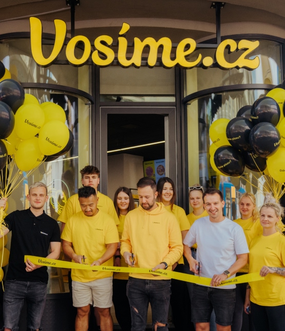 Group of people in yellow shirts participate in a ribbon-cutting outside the Vosime.cz storefront with yellow and black balloons, marking a grand opening.